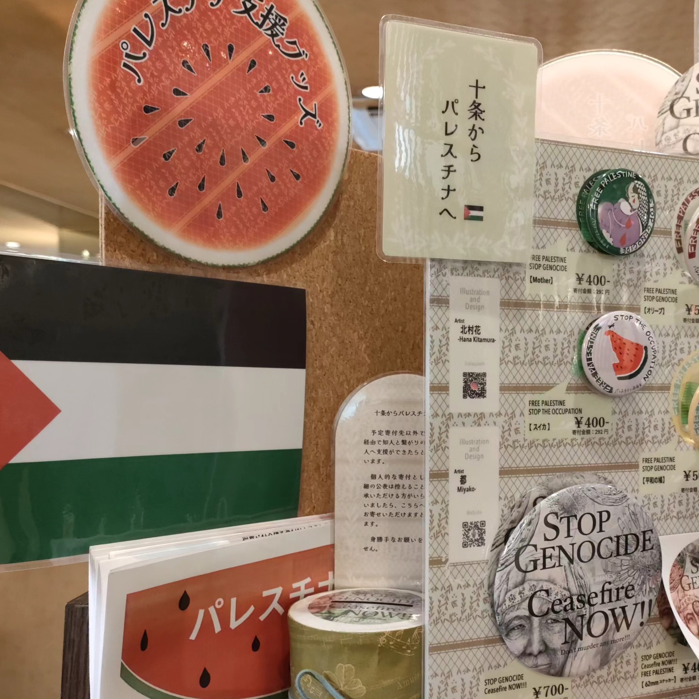 レジ横の物販スペース、パレスチナの旗や支援グッズ販売を周知するPOP、缶バッジ等の見本と価格を案内するパネルが並ぶところに、新たにチラシを置かせていただいた様子を移した写真です。