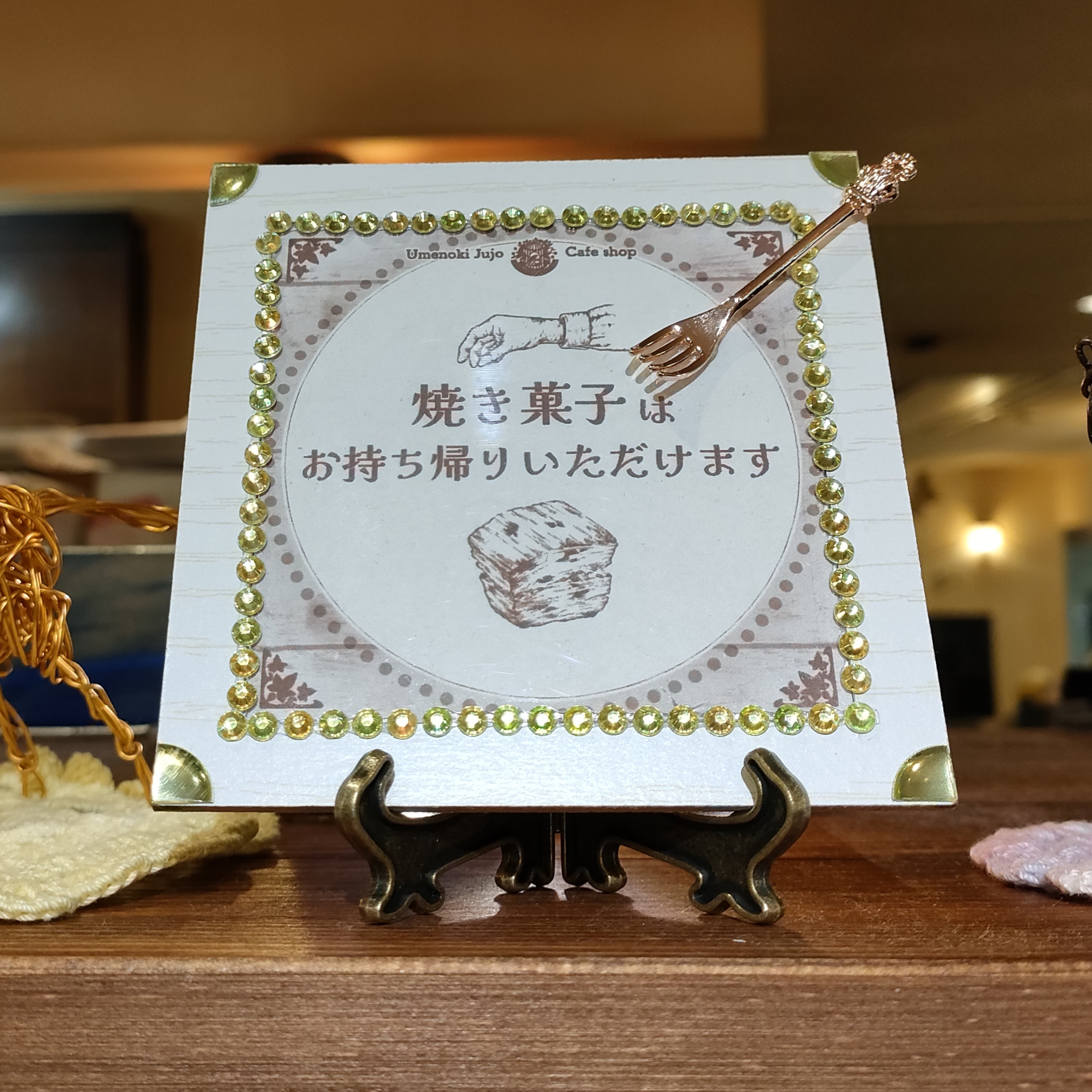 梅の木十条店さんの、焼き菓子お持ちかり可を宣伝するミニ看板制作風景です。端材の表面が白い木目の板をくり抜いてフレームの代用にし、ペールらいむぐりーんのらインストーンシールを枠フチに飾り、フレーム内にスコーンや手を描いたペン画イラストとテキストをレイアウトしています。フレームのコーナにはラインストーンシールと同色の光沢のある円形シールをカットして貼り、ミニチュアの金色のフォークを添えて雰囲気をあげています。